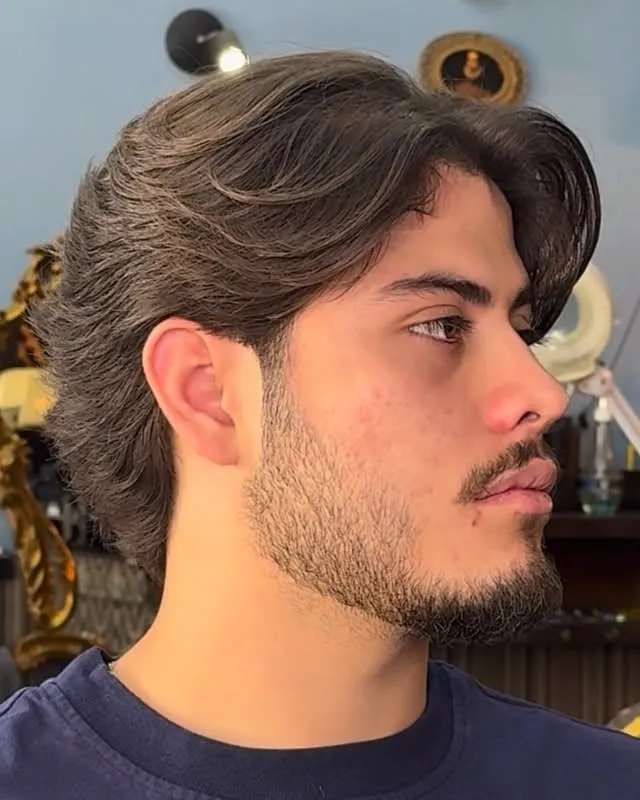 Hombre de perfil con corte bro flow de largo medio peinado hacia atrás, pelo detrás de las orejas y barba corta