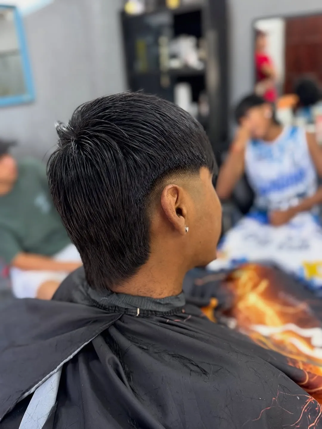 Joven de perfil en la barbería con burst fade bajo alrededor de la oreja, cabello negro lacio largo y nuca larga sobre el cuello
