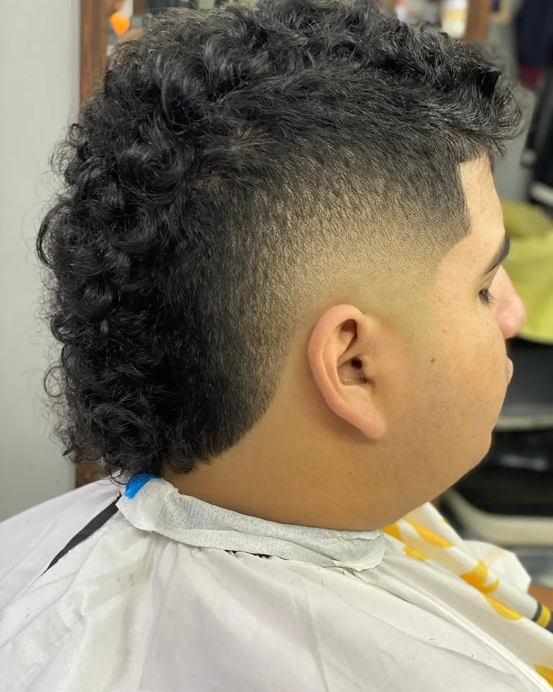 Joven de perfil con burst fade en cabello crespo negro, volumen arriba y nuca rizada larga