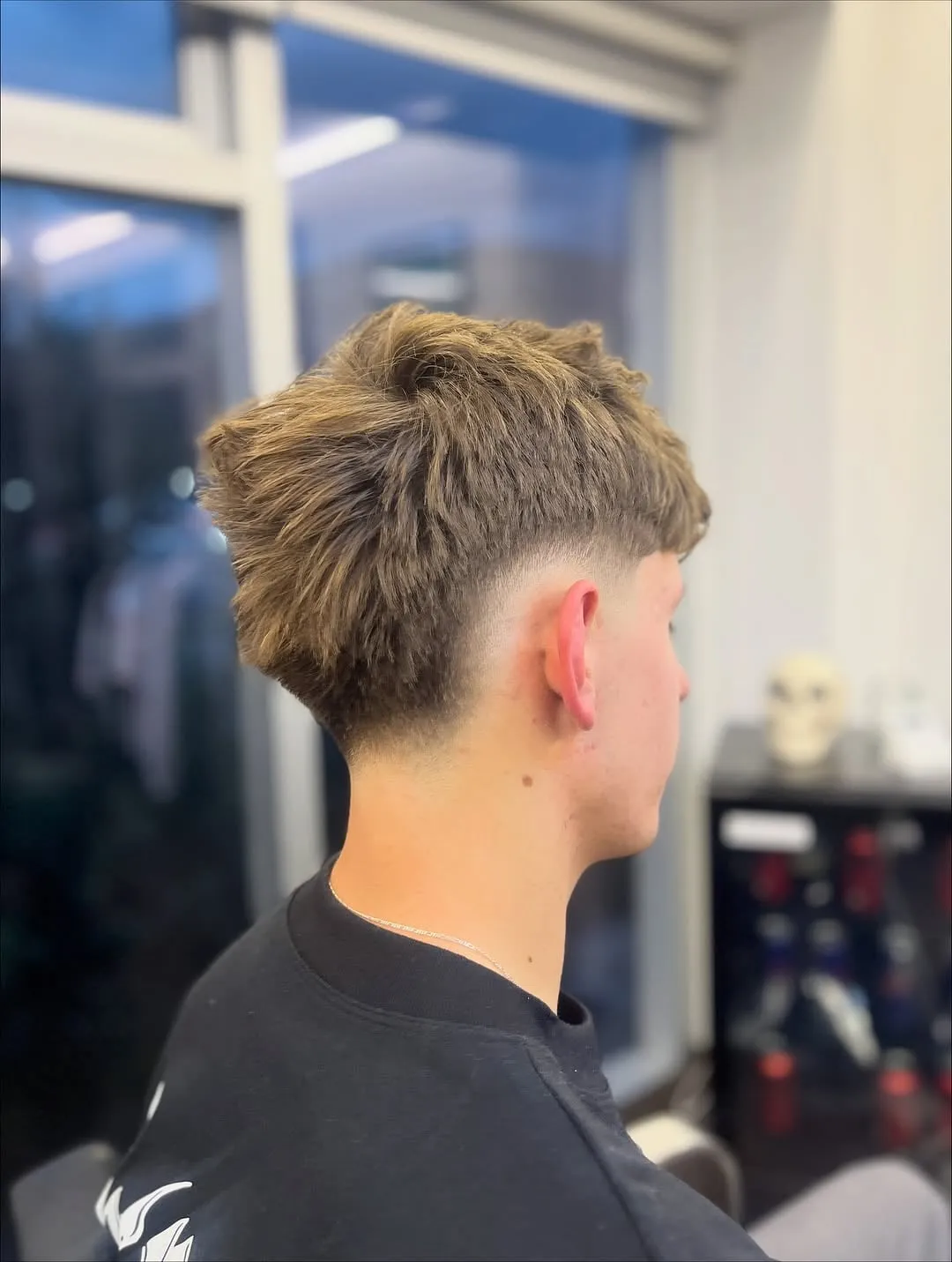 Joven visto de espaldas y de lado con burst fade bajo, cabello castaño claro texturizado arriba y nuca corta