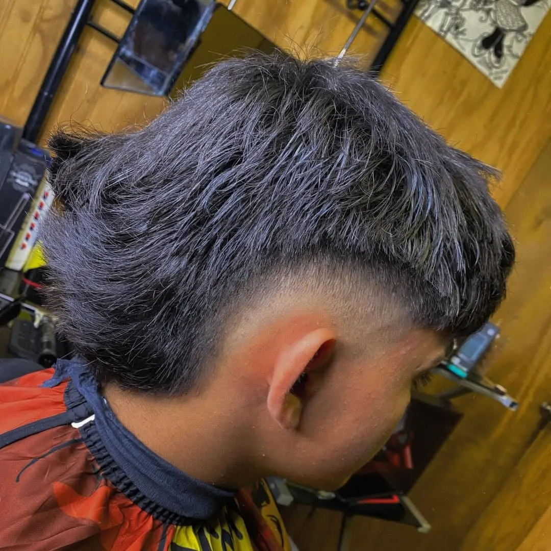 Hombre de lado con burst fade alrededor de la oreja y cabello negro con reflejos azulados, nuca terminada en cola de pato
