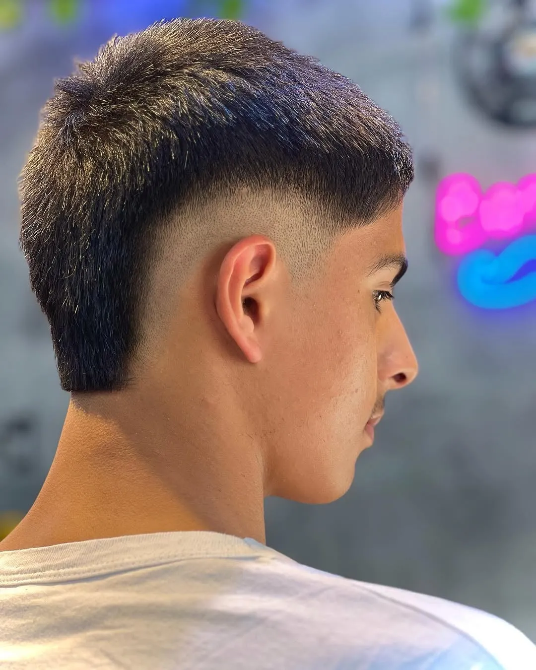 Joven de perfil con burst fade alto y cabello negro muy corto arriba, casi rapado con máquina
