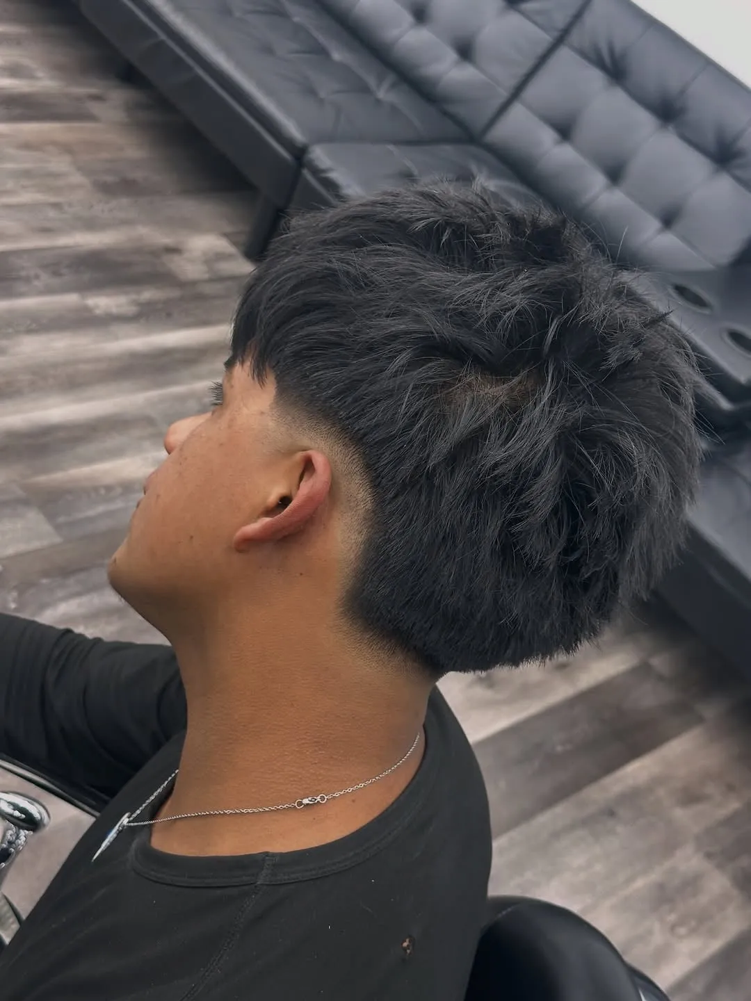 Joven de perfil con burst fade medio, desvanecido en curva sobre la oreja y cabello negro lacio despeinado hacia adelante