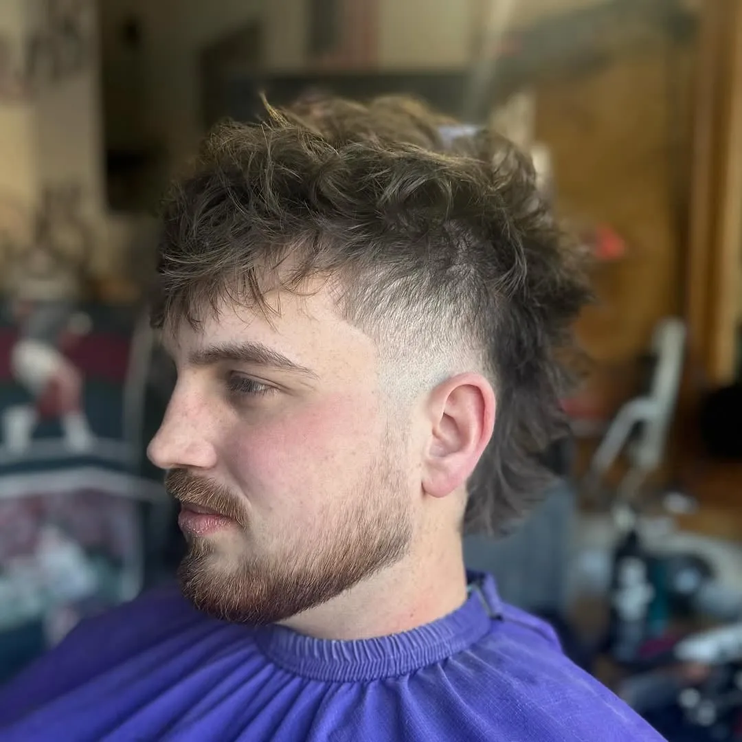 Hombre con barba y burst fade en semicírculo alrededor de la oreja, cabello castaño texturizado arriba y nuca larga