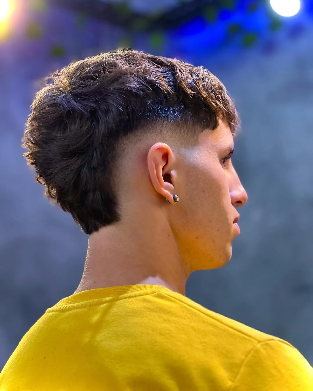 Joven de perfil con burst fade alrededor de la oreja y mullet de cabello castaño ondulado con nuca larga