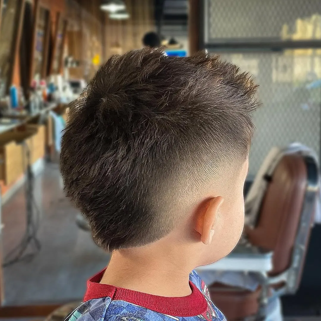 Niño visto de espaldas y de lado en la barbería con burst fade alrededor de la oreja y cabello castaño texturizado arriba