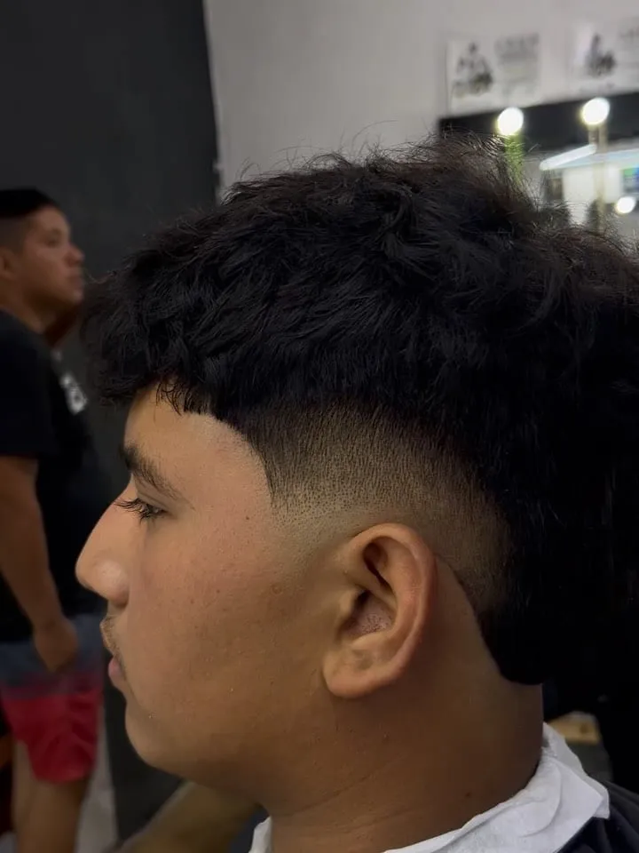 Joven de perfil con burst fade redondo que dibuja un semicírculo limpio alrededor de la oreja y cabello negro lacio arriba