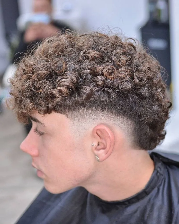 Joven de perfil con burst fade medio y rizos castaños abiertos que caen como fleco sobre la frente