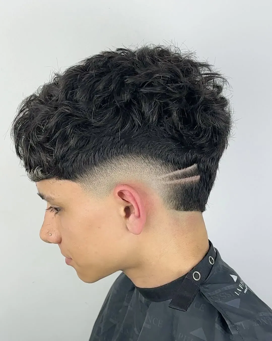 Joven de perfil con burst fade, rizos negros arriba y dos líneas paralelas dibujadas en diagonal sobre la oreja