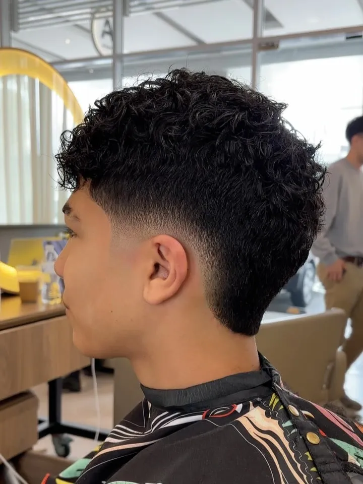 Joven de perfil con burst fade bajo y rizos cortos negros arriba, nuca rizada y patilla desvanecida