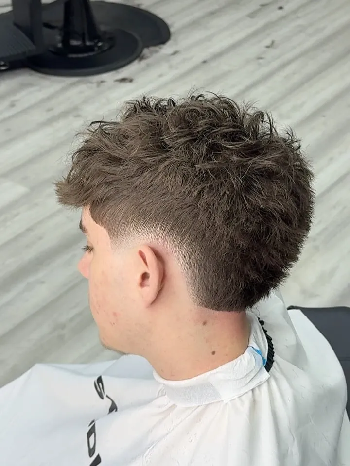 Joven visto de espaldas y de lado con burst fade bajo y cabello castaño ondulado texturizado arriba