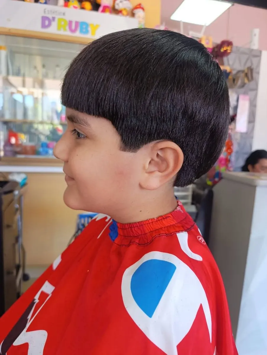 Niño de perfil con cabello negro lacio y brillante en forma de casquete, fleco recto y grueso hasta las cejas, corte horizontal sobre la oreja y taper corto en la nuca
