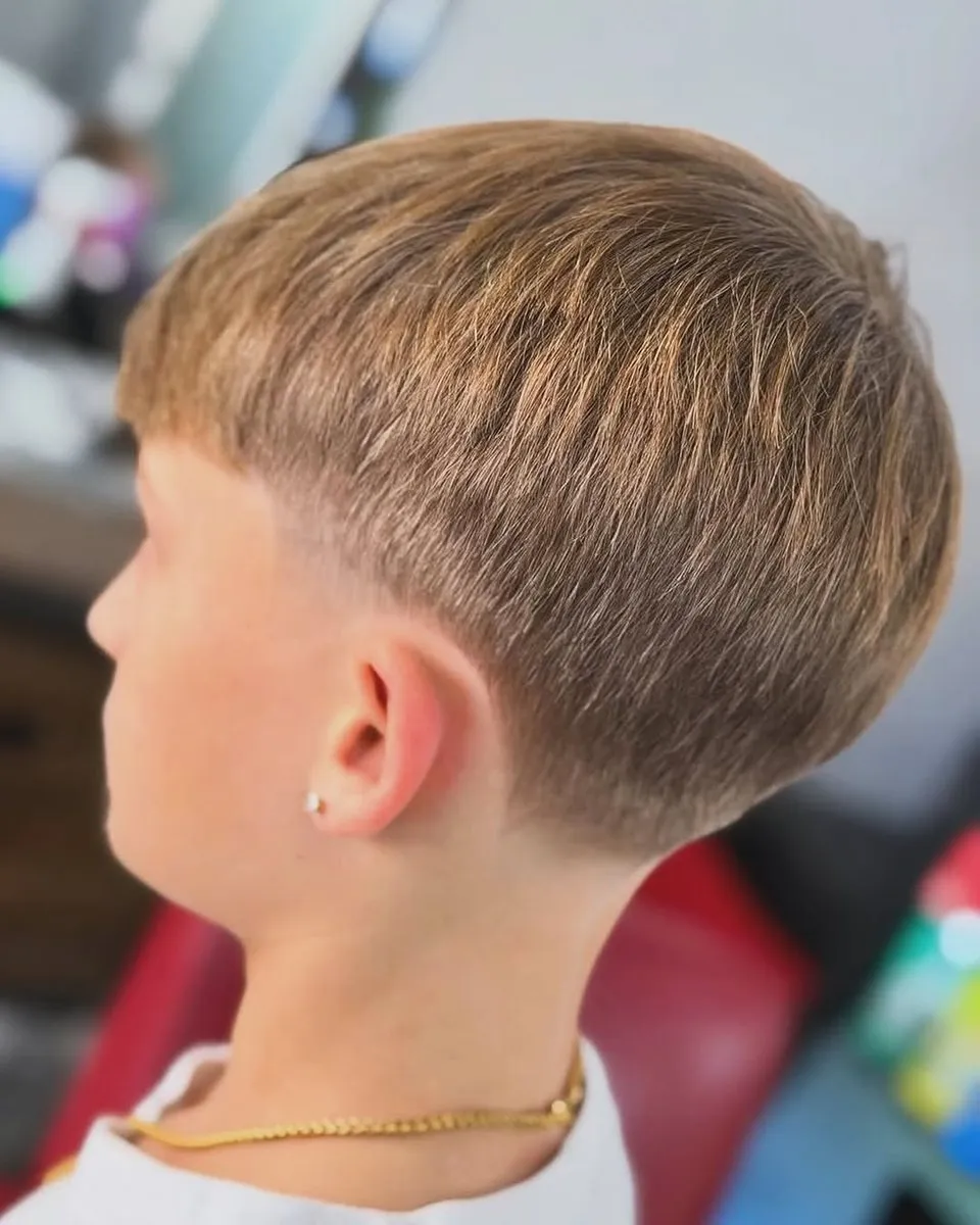 Niño visto de espaldas y de lado, con cabello rubio miel liso en forma de taza, fleco corto y recto y un taper bajo hasta la piel con límite definido bajo la taza