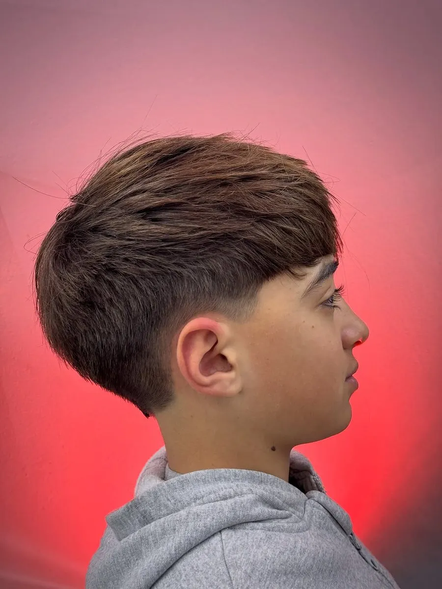 Adolescente de perfil sobre fondo rojo con cabello castaño texturizado, largo arriba y cayendo hacia adelante en un fleco desfilado, con low a mid fade sobre la oreja y límite suave