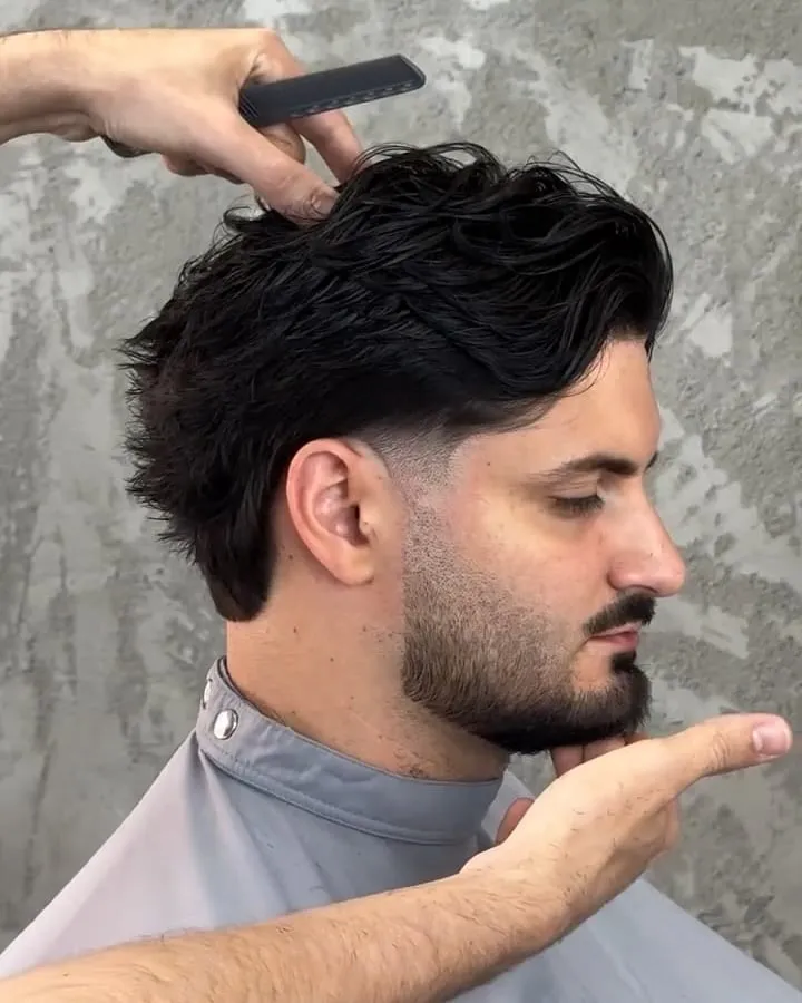 Hombre con corte medio texturizado ondulado, taper suave y barba recortada mientras el barbero lo peina