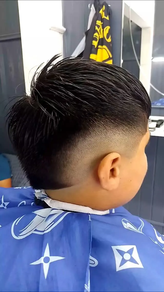 Niño visto de espaldas y de lado con cabello negro liso largo arriba, de unos 6 a 7 cm, peinado hacia atrás y hacia arriba en una cresta, y fade a piel que rodea la oreja en arco
