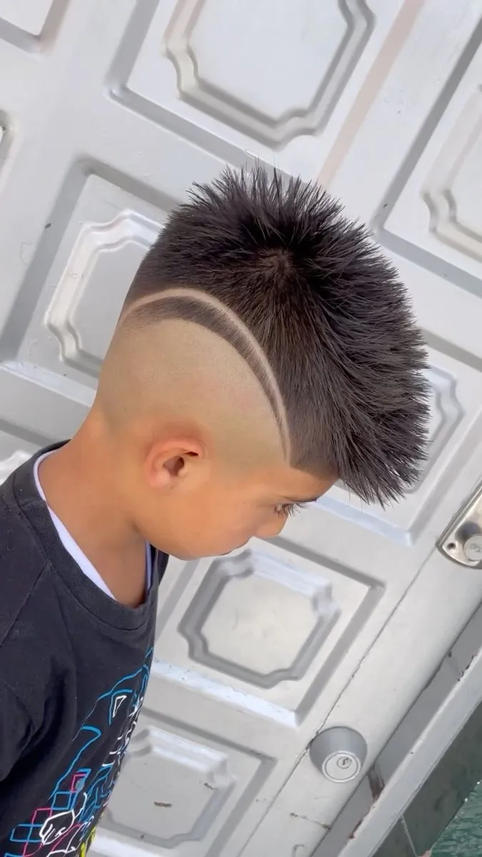 Niño visto de lado y desde arriba, mirando hacia abajo, con cabello negro en el centro levantado en puntas con gel, costados con high fade casi a piel y una línea curva del lado que va de la sien a la nuca