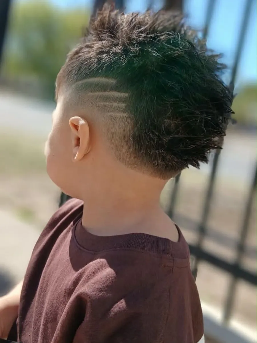 Niño al aire libre visto de espaldas y de lado, con cabello castaño oscuro en el centro revuelto en puntas, mid fade en el costado y la nuca, y cuatro rayas horizontales cortas y paralelas en el costado