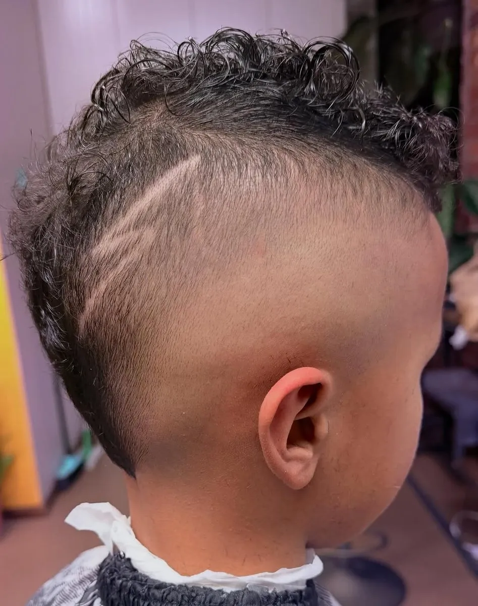 Primer plano lateral de un niño con una franja ancha de rizos negros pequeños en el centro de la cabeza, skin fade a piel en el costado y dos líneas curvas paralelas sobre la oreja