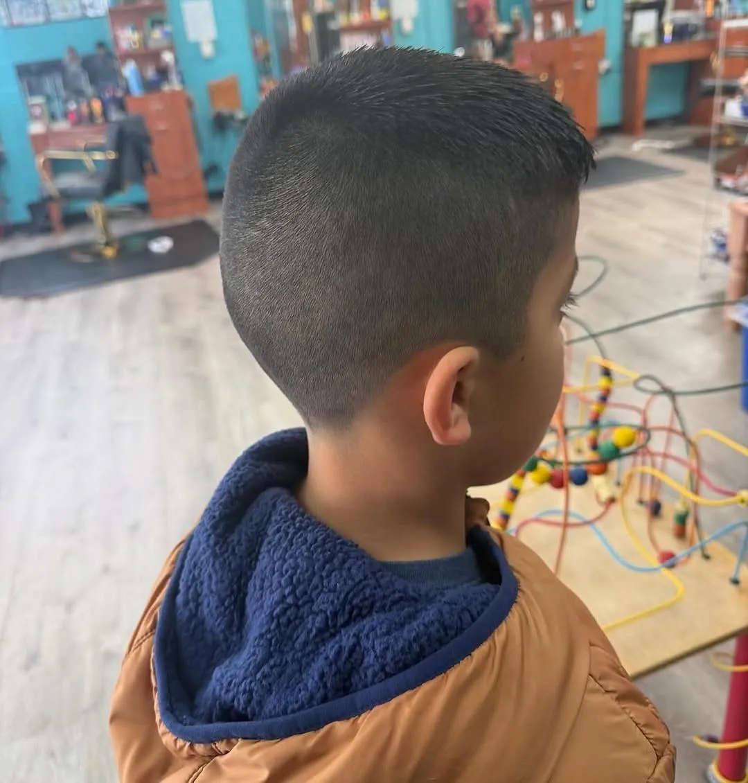 Niño de espaldas y de lado en una peluquería infantil, con cabello oscuro igual de corto en toda la cabeza, de unos 1 cm arriba y apenas más corto hacia el cuello, sin dibujos ni líneas
