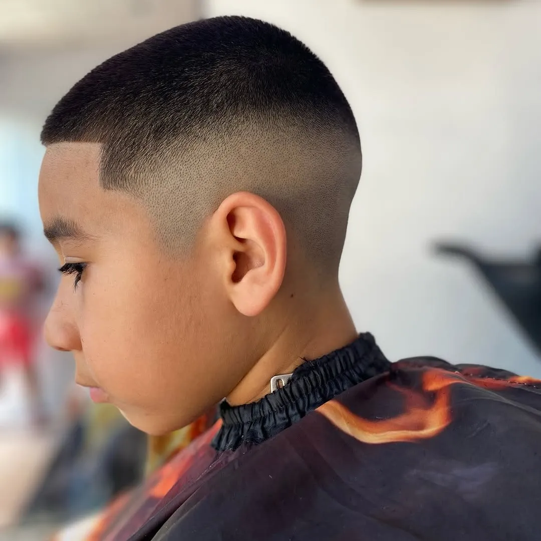 Niño de perfil con cabello negro de 0.5 a 1 cm arriba, high fade a piel en el costado y la nuca y contorno recto en la frente con ángulo en la sien
