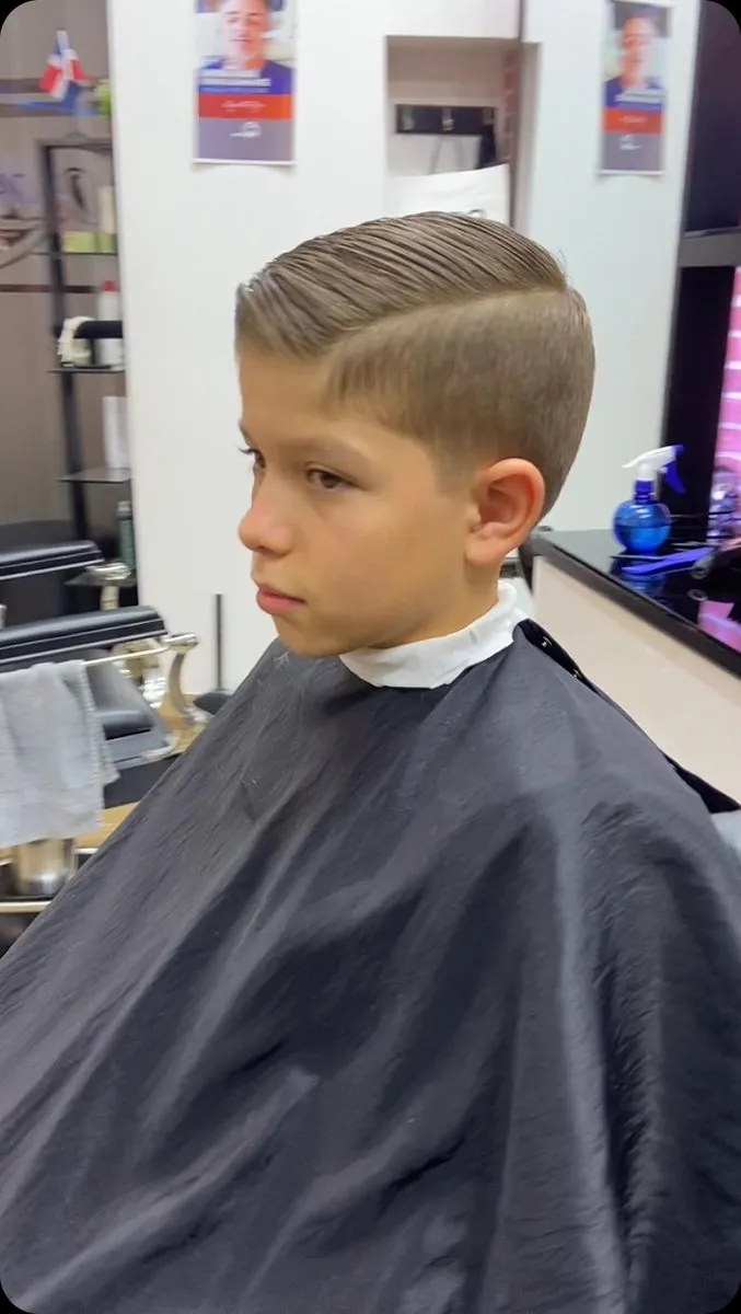 Niño en tres cuartos de perfil en una barbería, con cabello castaño claro y liso de 4 a 5 cm peinado liso hacia un lado con raya, low fade junto a la oreja y sienes rectas
