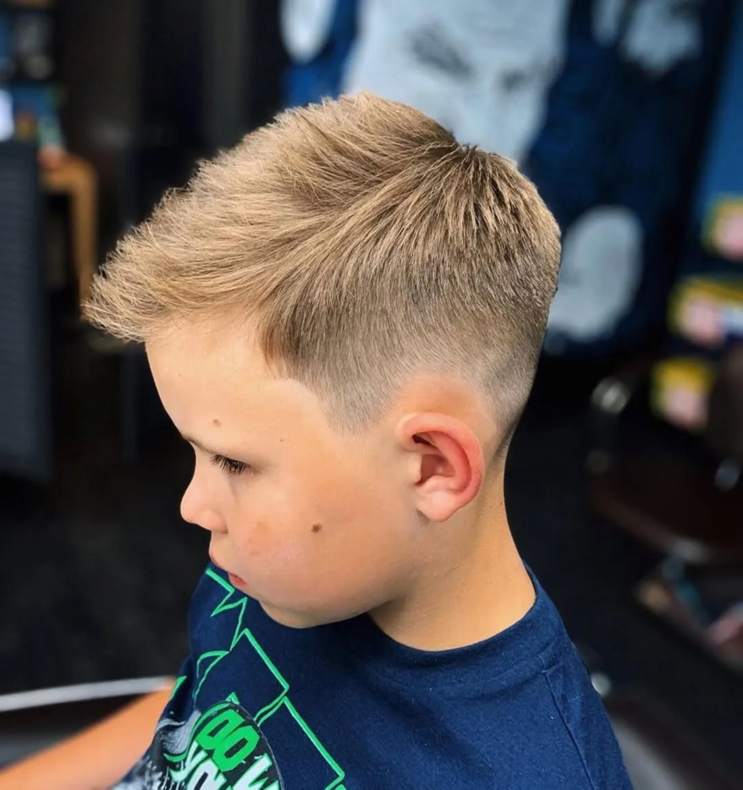 Niño de perfil con cabello rubio claro de 4 a 5 cm texturizado en mechones hacia adelante y arriba, mid fade que deja ver la piel sobre la oreja y contorno recto en la sien