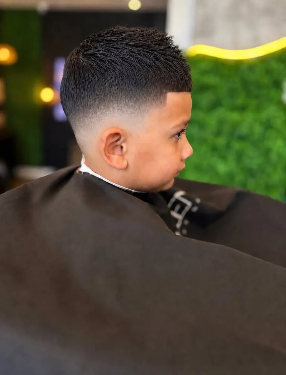 Niño de perfil con cabello negro de 2 a 3 cm levantado hacia adelante, mid fade hasta la piel junto a la oreja y la frente y la sien marcadas con una línea recta