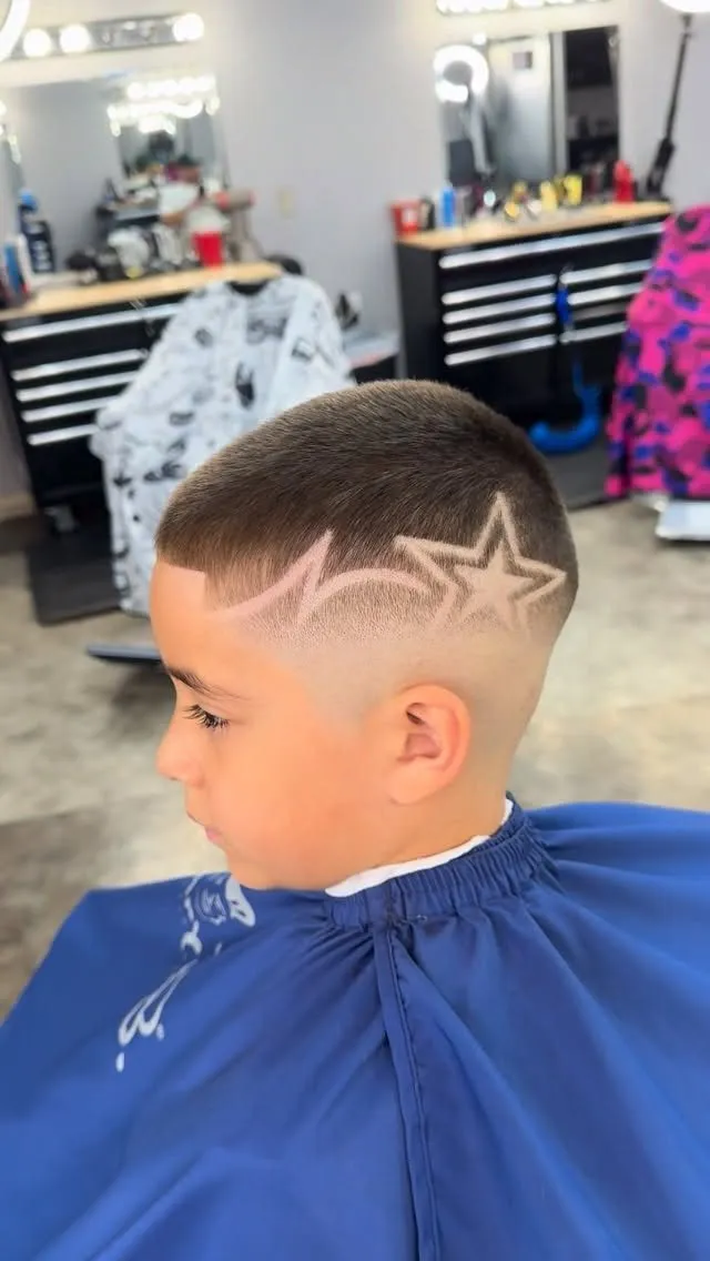 Niño de perfil en una barbería con capa azul, cabello de 1 cm arriba, high fade a piel y una estrella con cola en zigzag, como una estrella fugaz, rapada sobre la oreja