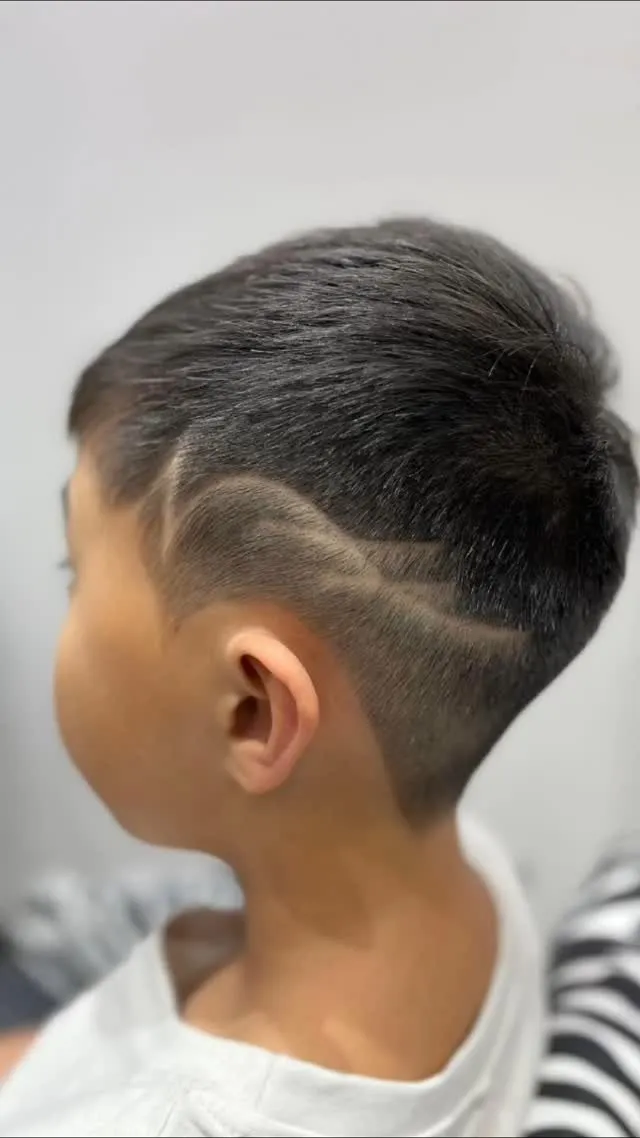 Niño de espaldas y de lado con cabello negro liso corto arriba y peinado hacia atrás, taper bajo a piel detrás de la oreja y una línea ondulada suave que va de la sien a la nuca