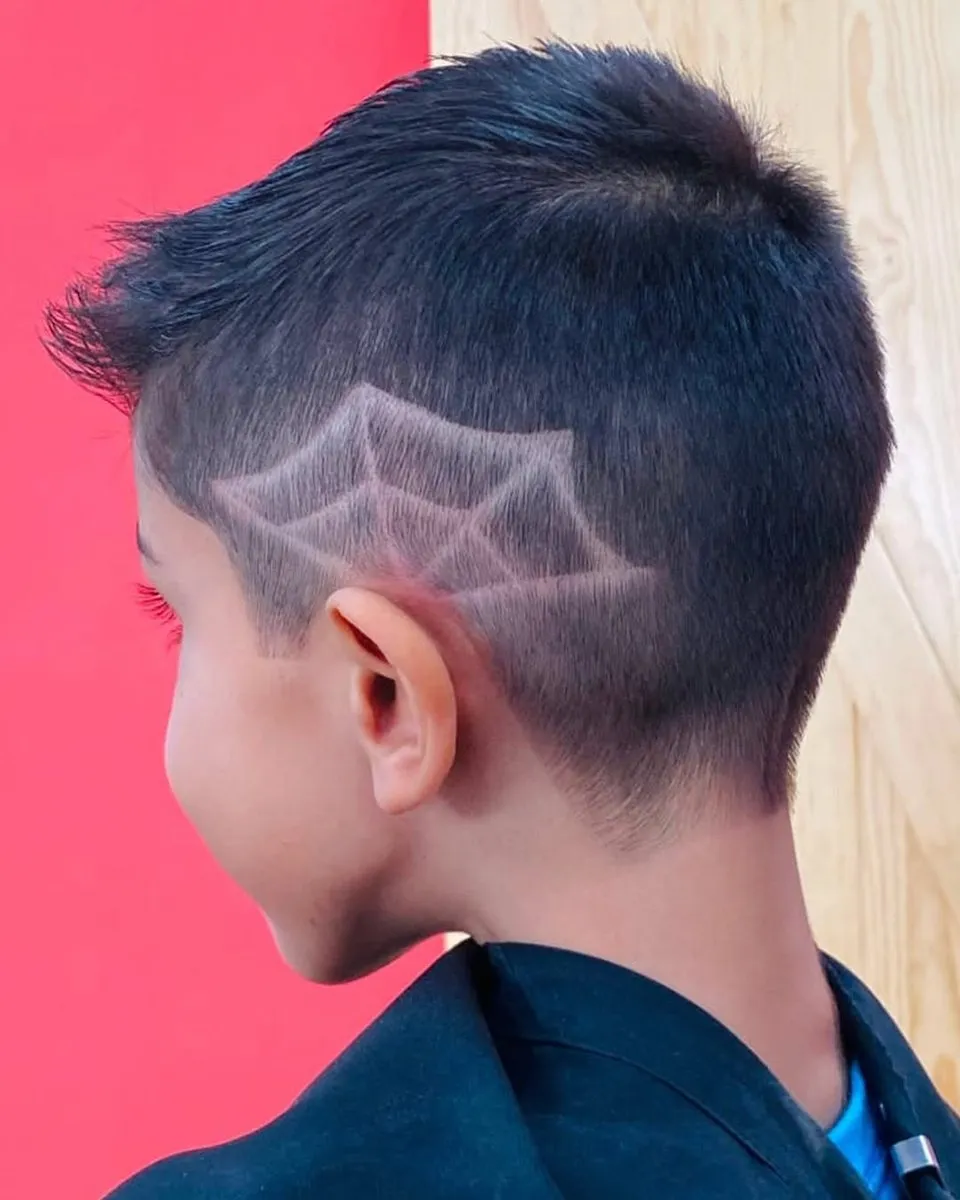 Niño visto de espaldas y de lado con cabello negro corto arriba, de unos 3 cm, llevado hacia adelante, low a mid fade y una telaraña con rayos y arcos rapada sobre la oreja