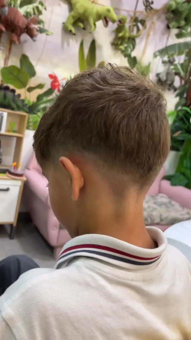 Niño visto de espaldas y de lado en una peluquería infantil, con cabello castaño claro texturizado arriba, low fade y un pico en la nuca que termina en ángulo hacia abajo