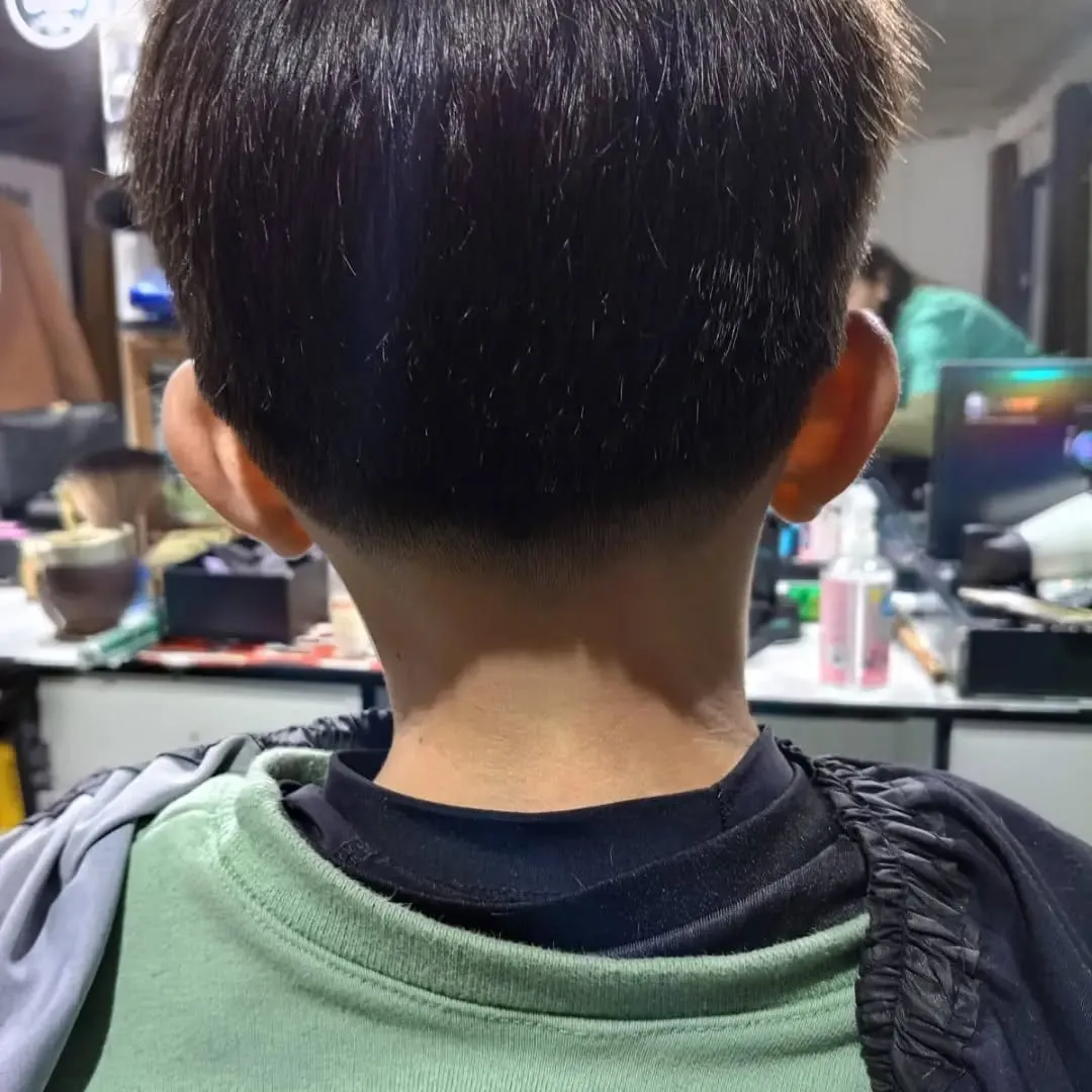 Niño de espaldas con cabello negro lacio de 3 a 4 cm que cae hacia abajo y un taper bajo que se pierde en la nuca, con las orejas despejadas