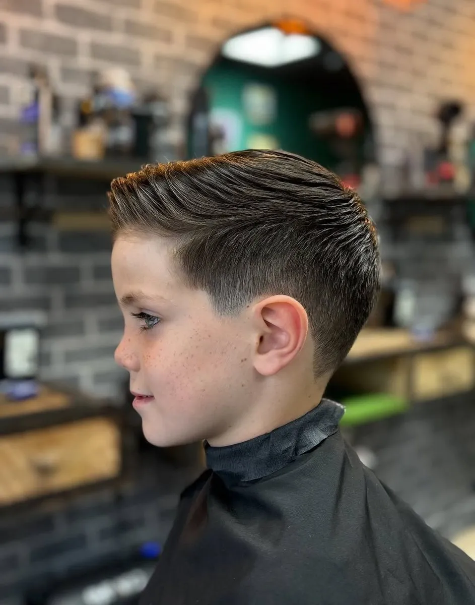Niño de perfil en una barbería con pared de ladrillo, cabello castaño de 5 a 6 cm peinado hacia atrás con textura, low taper en la sien y detrás de la oreja, lados cortos sin llegar a piel y contorno suave en la frente
