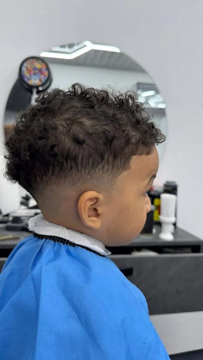 Niño pequeño de lado y de espaldas con capa azul clara, rizos castaño oscuro de 3 a 4 cm arriba, low a mid fade sobre la oreja y en la nuca y contorno suave en la frente