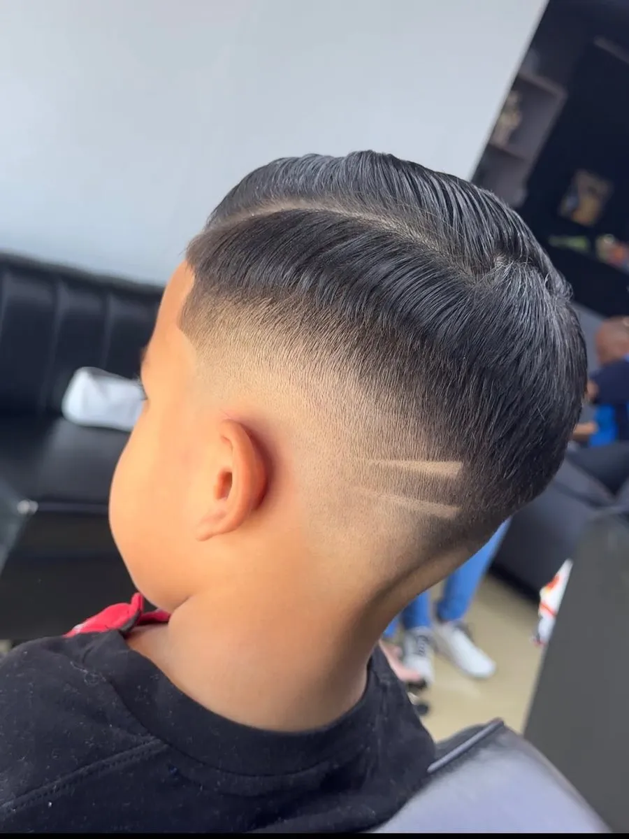 Niño de espaldas y de lado con cabello negro liso de unos 5 cm peinado liso hacia un lado, una raya rapada de la sien a la nuca, mid fade a piel y tres rayas oblicuas paralelas en la parte baja de la nuca