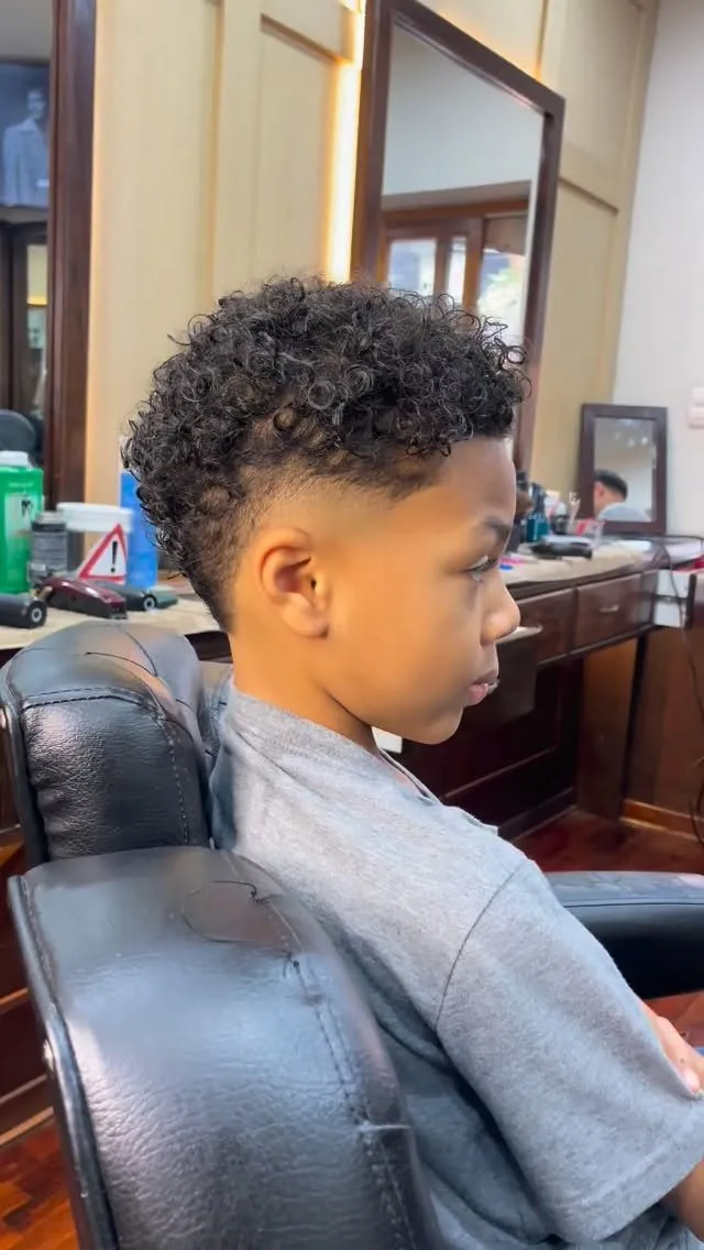 Niño de perfil sentado en el sillón, con rizos negros pequeños de 4 a 5 cm de volumen arriba que caen un poco sobre la frente, mid fade a piel en arco alrededor de la oreja y nuca corta