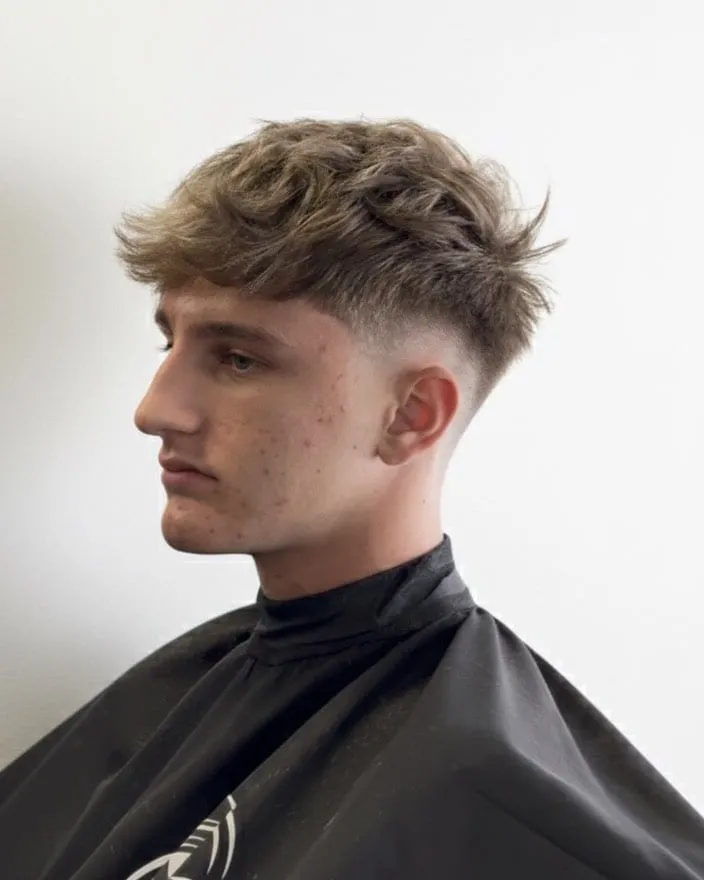 Joven de perfil con fleco texturizado rubio cenizo que cae hacia la frente y low taper fade
