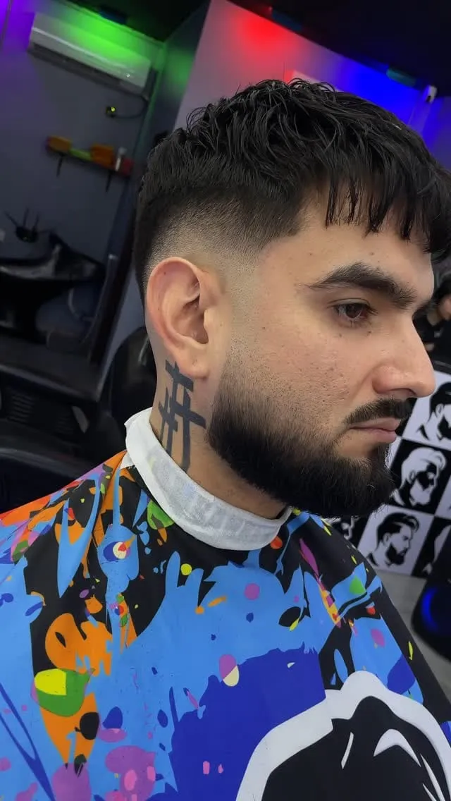 Hombre de perfil con barba completa, french crop negro con fleco recto texturizado y mid fade conectado a la barba