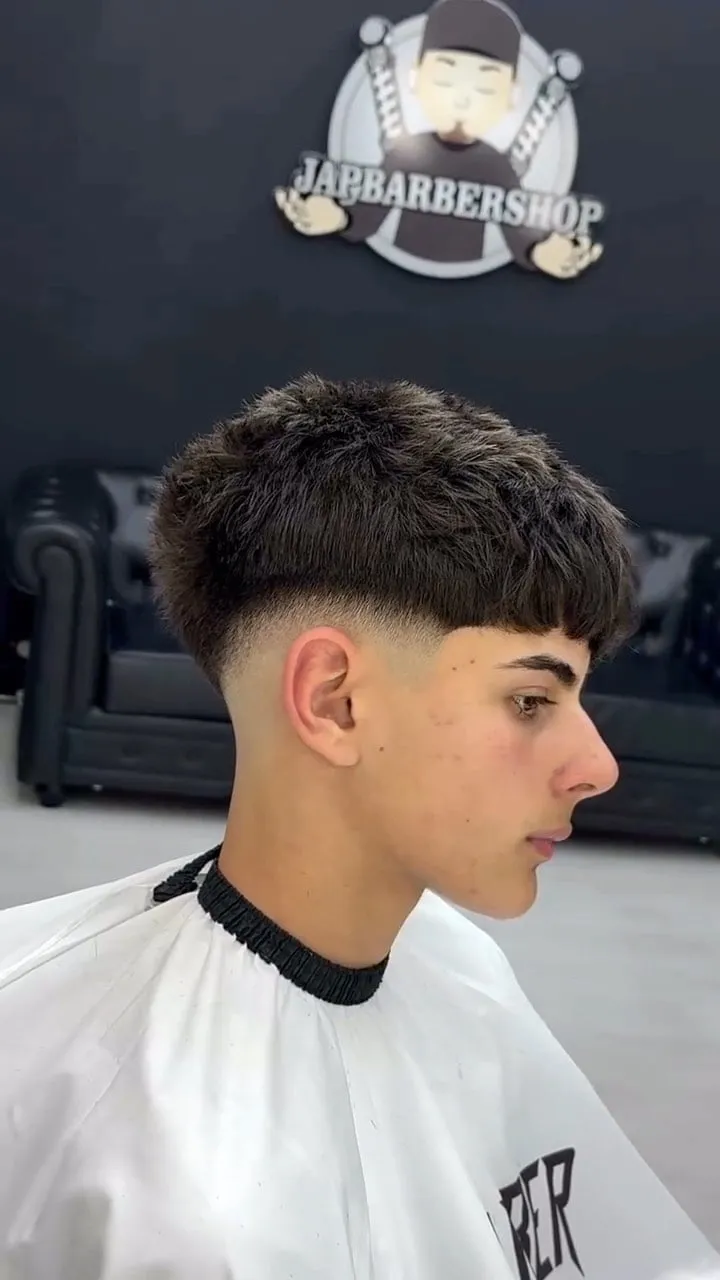 Adolescente de perfil con french crop negro, fleco recto marcado sobre las cejas y low fade que deja ver la piel en la sien