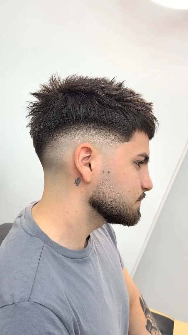 Hombre de perfil con barba corta, french crop negro con puntas hacia adelante y high fade que llega a la sien