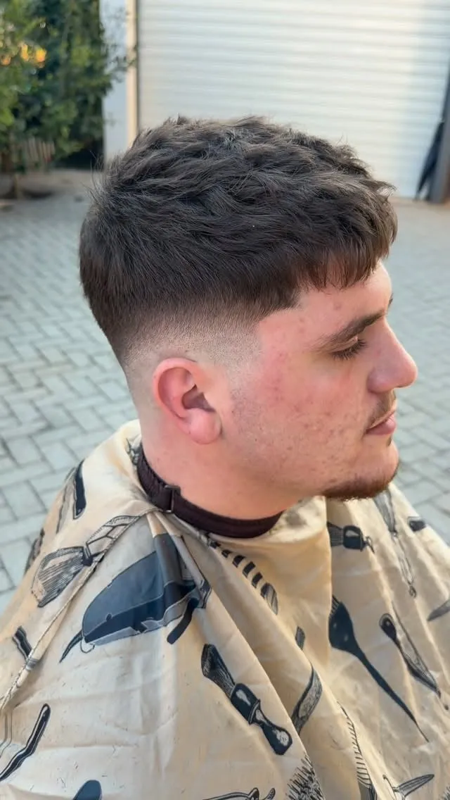 Joven de perfil con french crop castaño oscuro, textura arriba, fleco corto irregular y mid fade que sube hasta la sien