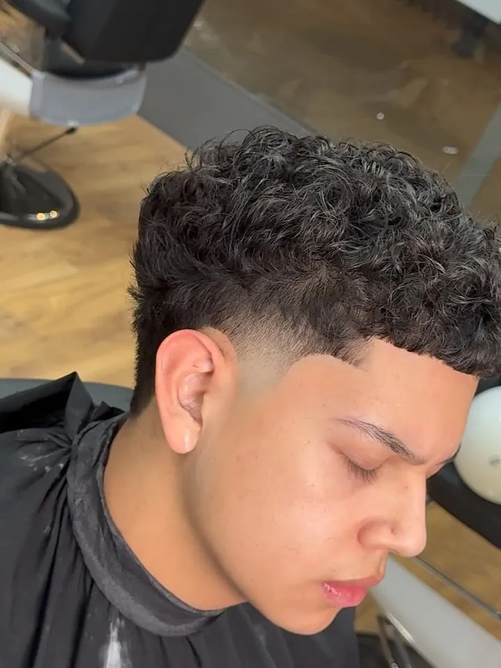Joven de perfil con french crop en rizos negros pequeños, fleco rizado sobre la frente y taper bajo en la sien