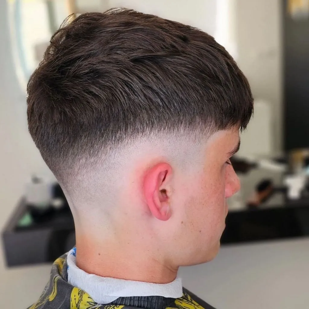 Joven visto de perfil y algo de espaldas con french crop castaño oscuro texturizado y skin fade que baja a la piel alrededor de la oreja