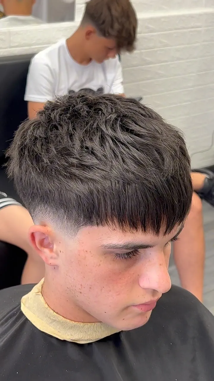Joven en la barbería con french crop texturizado castaño oscuro, fleco recto grueso a la altura de las cejas y low fade sobre la oreja
