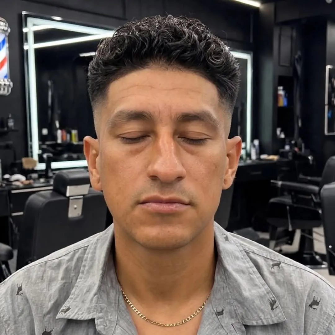 Hombre de frente en una barbería con cabello rizado corto arriba y taper fade en las patillas