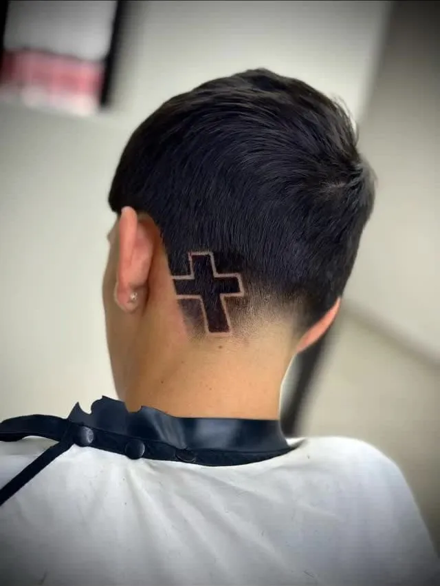 Taper fade bajo visto de espaldas con una cruz rellena dibujada junto a la oreja en cabello negro lacio