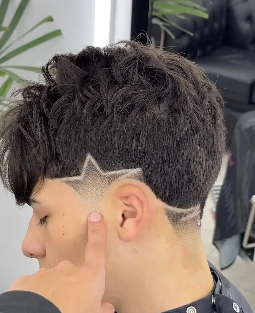 Taper fade de perfil con una estrella dibujada sobre la oreja y una línea que baja hasta la nuca en cabello castaño oscuro texturizado