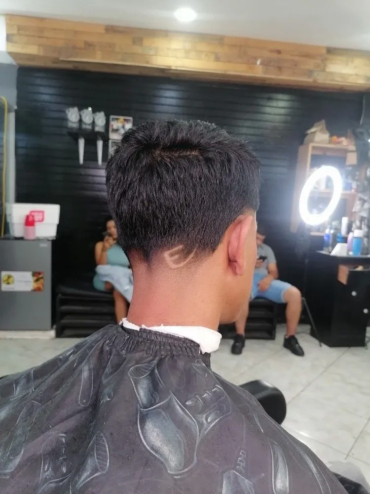 Taper fade bajo visto de espaldas con una letra E pequeña dibujada detrás de la oreja en cabello negro corto