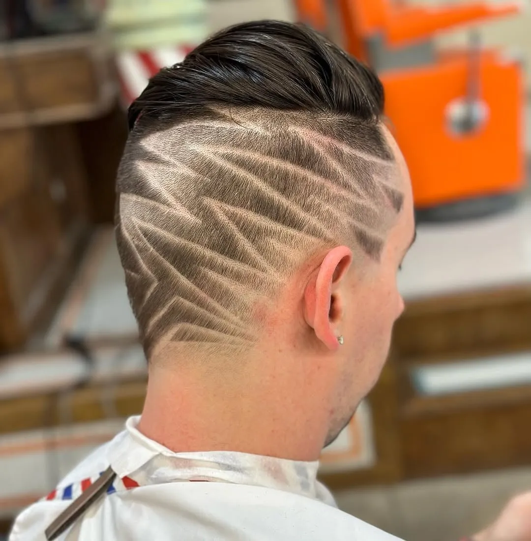 Corte con desvanecido alto y líneas en zigzag que cubren todo el costado de la cabeza con cabello peinado hacia atrás